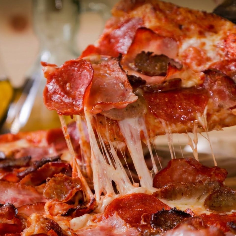 Close-up of a meat-lovers pizza slice lifted from a cheesy pie, showing pepperoni, sausage, ham, and stretchy melted cheese.