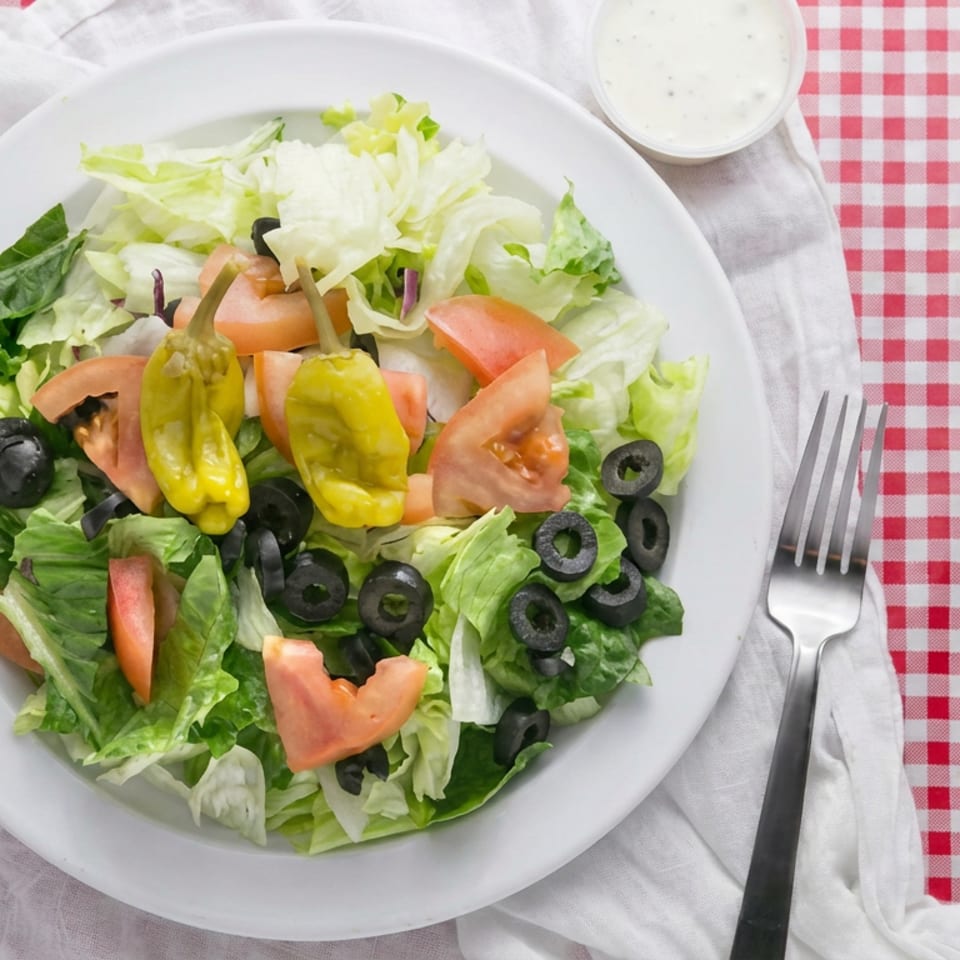 Fresh garden salad with lettuce, tomatoes, black olives, pepperoncini, and dressing on a red-and-white checkered tablecloth.
