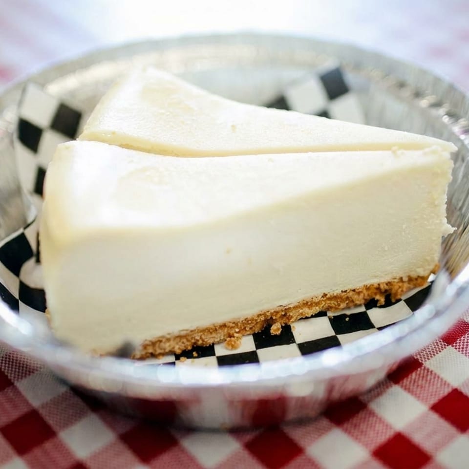 Two slices of creamy cheesecake with graham cracker crust in a foil dish on a red-and-white checkered tablecloth.