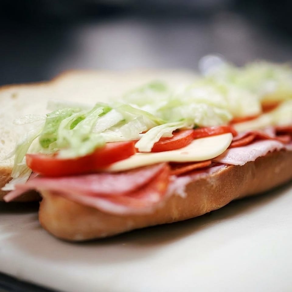 Open-faced sandwich layered with ham, salami, tomato, lettuce, and mayonnaise on a toasted roll.