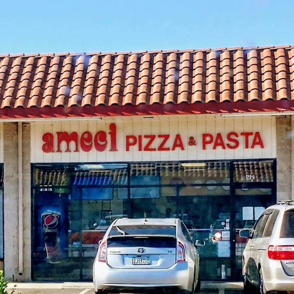 Ameci Pizza & Pasta storefront with terracotta tile roof, glass entrance, parked cars, and neighboring salon signage.