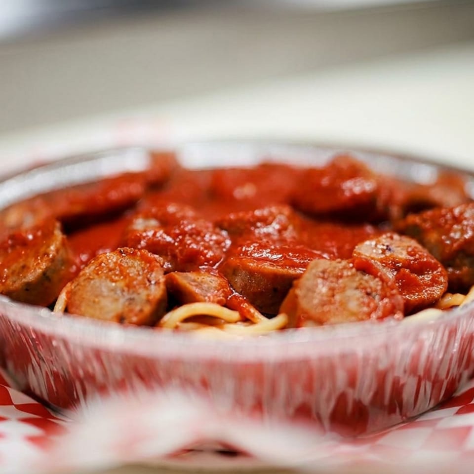 Spaghetti topped with sliced sausage and meatballs in tomato sauce, served in a foil bowl on red-and-white checkered paper.